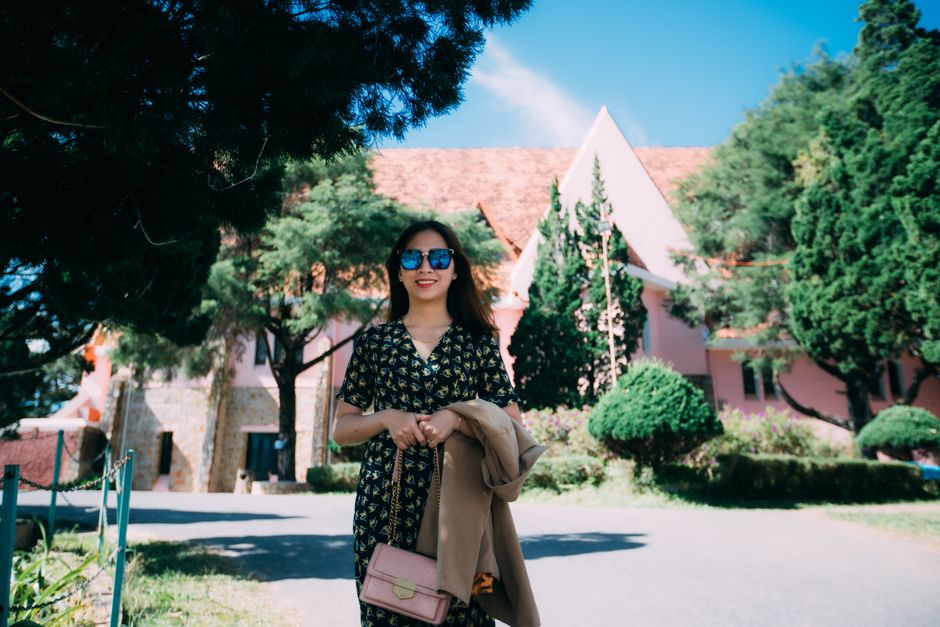 Woman in sunglasses smiling outside a pink building under clear blue sky.