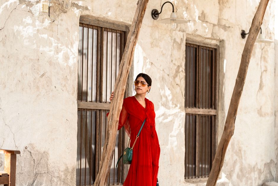 A fashionable woman in a red dress poses by a rustic wall in Dubai, UAE.