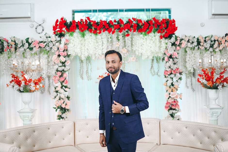 A portrait of a groom in formal attire, surrounded by vibrant floral decorations at an indoor wedding.