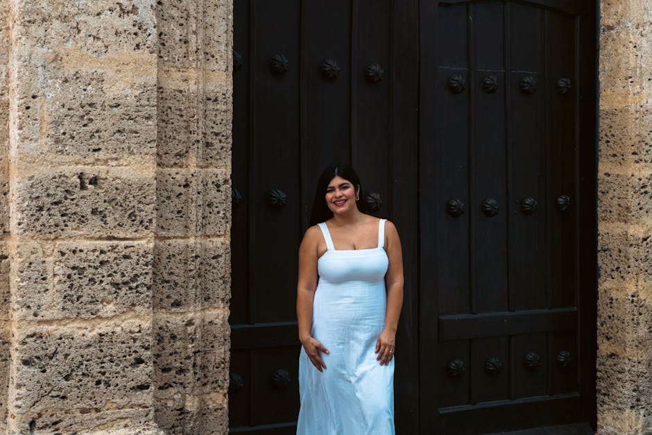 Woman in a white dress smiling by an ornate wooden door in a historic building.