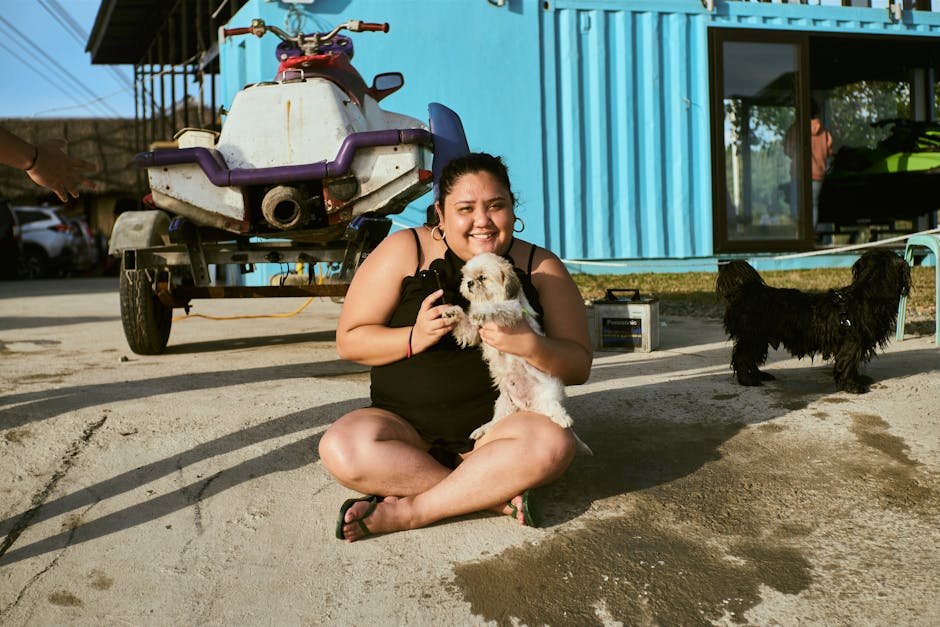 A woman in a black tank top holds two dogs, sitting in front of a colorful building.