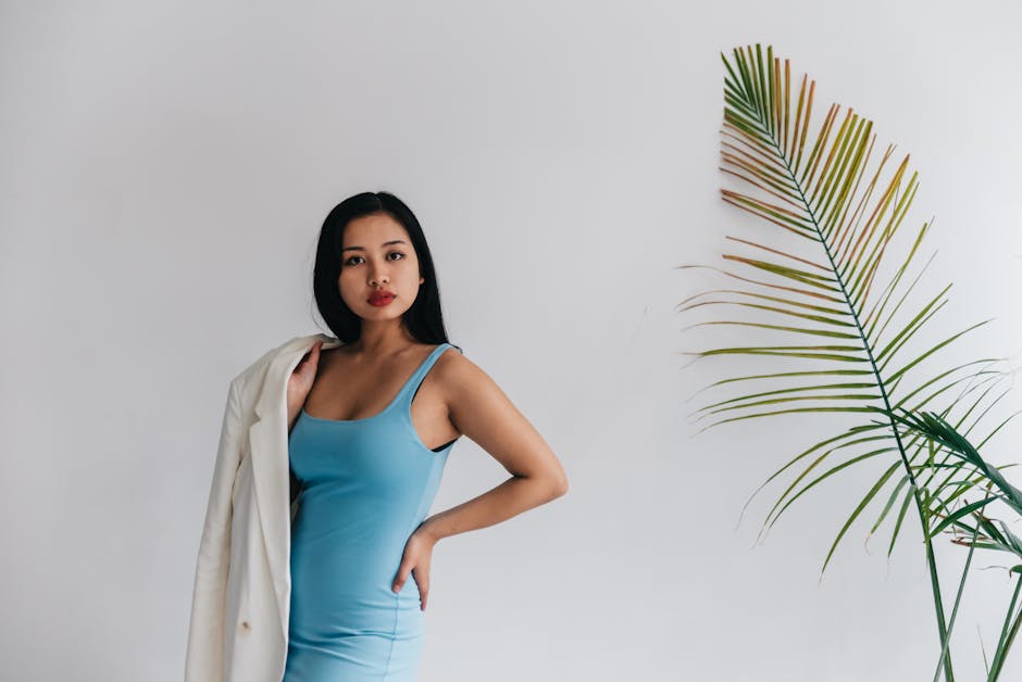 Elegant portrait of an Asian woman in a blue dress with white coat, posing by a minimalist indoor plant.