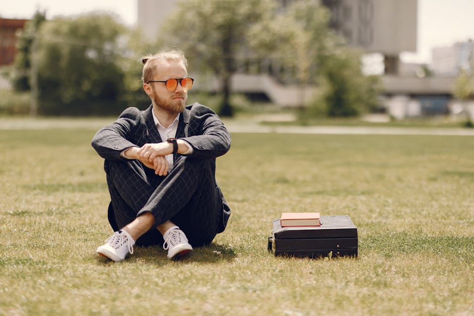 A man in a suit sits on grass in a park, wearing sunglasses and next to a briefcase with a book on top.