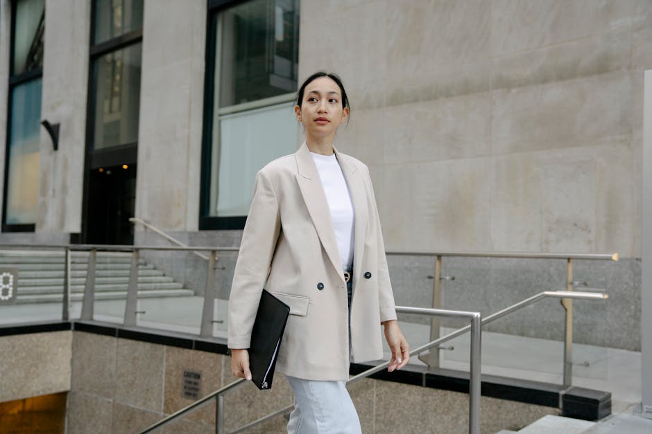 Confident Asian female entrepreneur in stylish outfit and with folder walking in city street and looking away