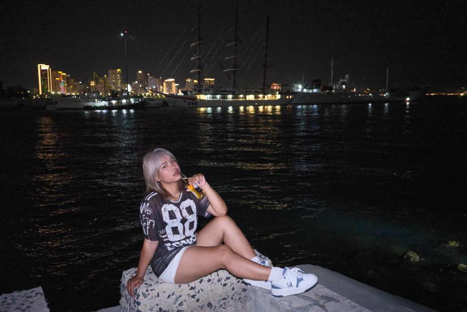 A young woman relaxes by the waterfront at night with a view of city lights and boats.