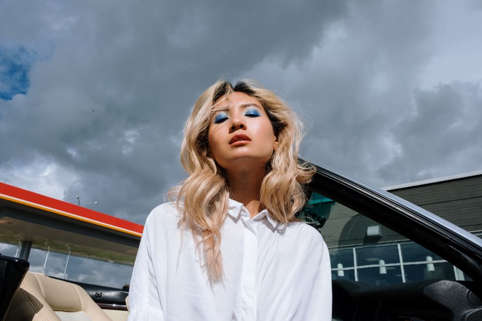 Portrait of a fashionable woman with blue eyeshadow standing by a car under a cloudy sky.