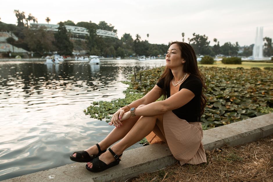 A woman peacefully sits by the lakeside in a park, enjoying the scenic view and serene atmosphere.