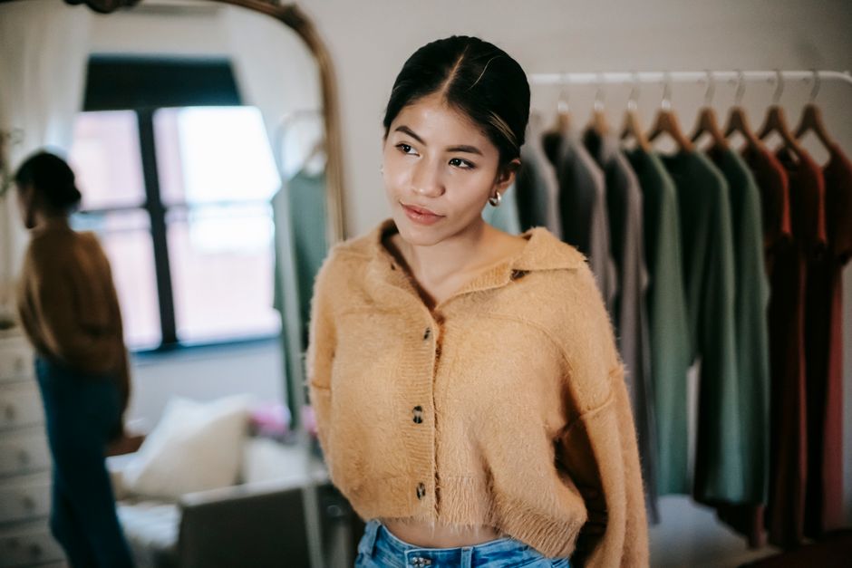 Thoughtful ethnic female in casual stylish clothes standing near mirror and rail with clothes in apartment