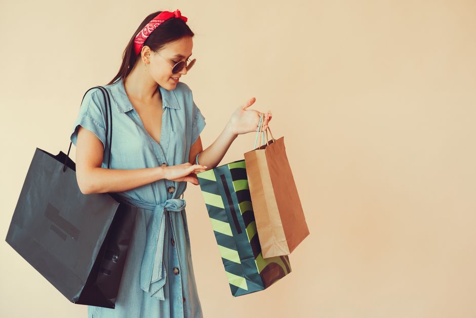 Cheerful young woman in trendy sunglasses and red headband looking into paper bags and holding shopping bag on shoulder during shopping on beige background