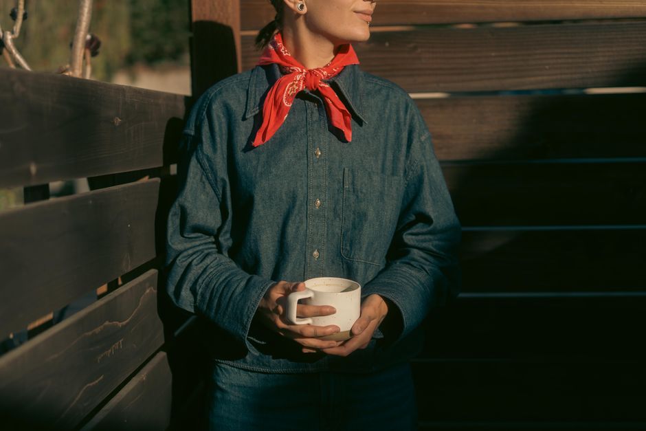 Person in denim shirt holding coffee cup, wearing red bandana. Outdoors, relaxed vibe.