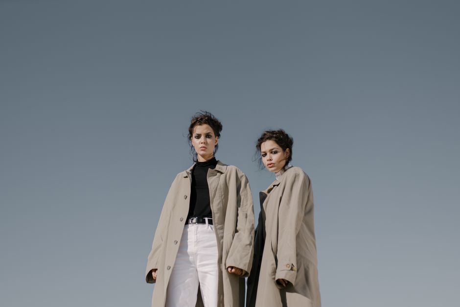 Two women in trench coats posing confidently against a clear sky.