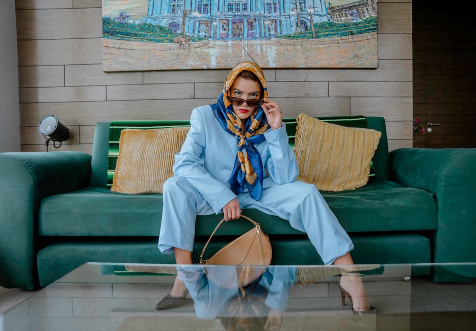 Fashionable woman in a blue suit with a scarf indoors, seated on a green couch with artwork backdrop.