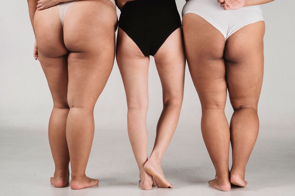 Back view lower body part of slim woman in black bodysuit standing between plus size females in bikini against white background in studio