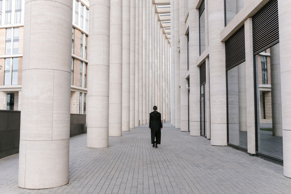 A person in black attire walks through a modern hallway with tall columns and reflective glass.