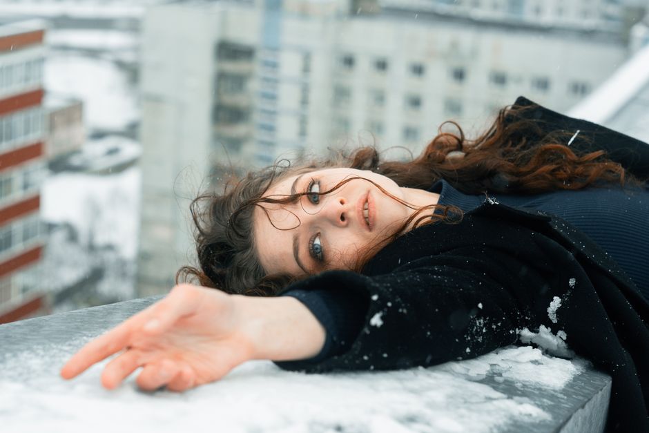 A woman lying on a snowy rooftop with an urban background, displaying artistic expression.