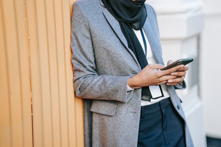 Crop unrecognizable female manager in stylish elegant outfit and name tag leaning on corrugated metal wall on street and text messaging on smartphone