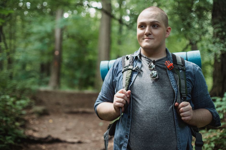 Smiling plus size male backpacker standing on path in green dense forest during hiking
