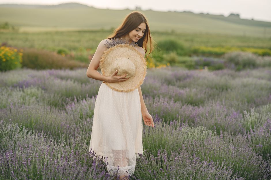 A woman wearing a white skirt enjoys a lavender field, smiling and holding a straw hat.