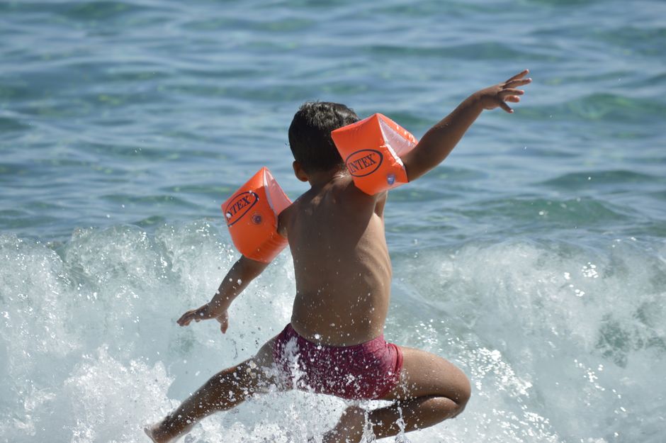A child wearing orange floaties joyfully jumps through ocean waves on a sunny day.