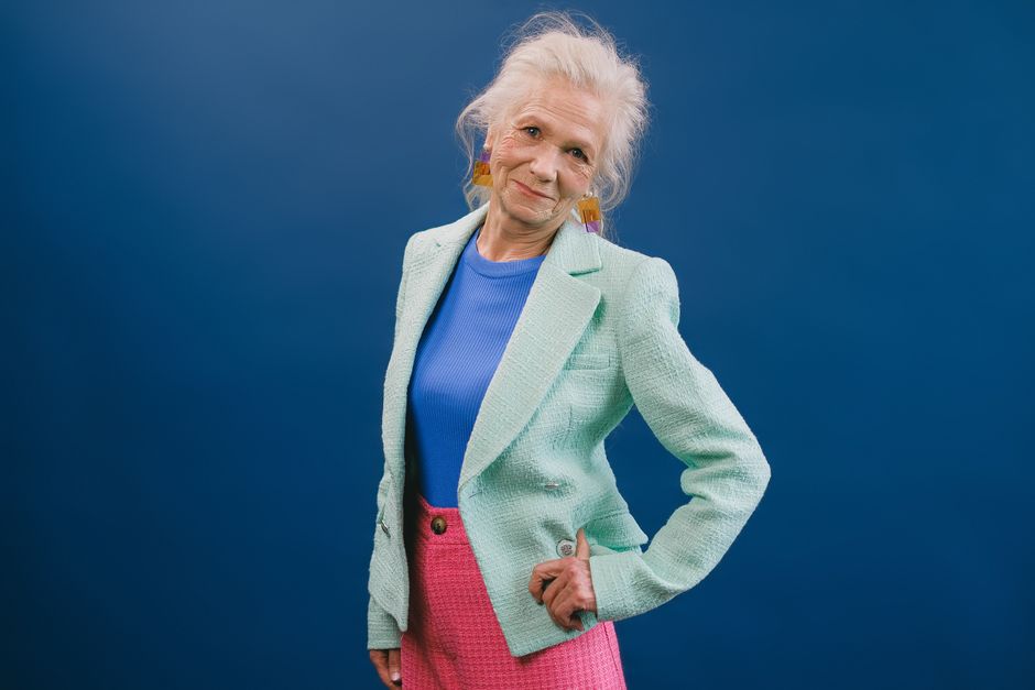 Delightful gray haired female in stylish green suit putting hand on waist and looking at camera against blue background