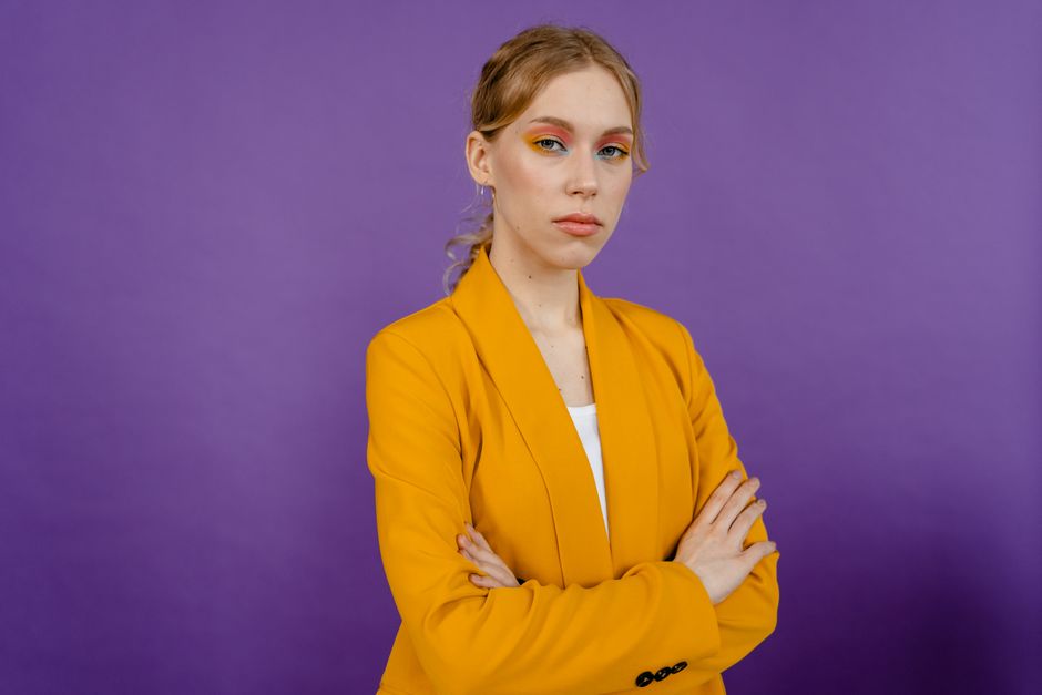 Portrait of a confident woman in a yellow blazer with colorful eye makeup against a purple background.