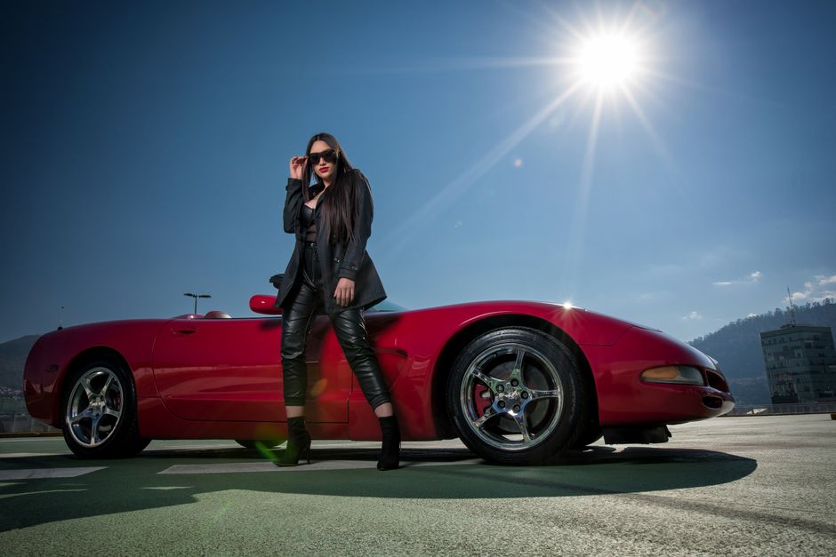 A fashionable woman poses confidently next to a sleek red sports car under the bright sun.