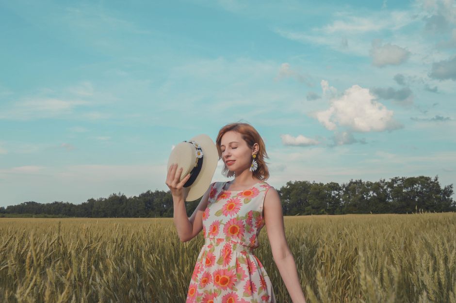 Positive female with closed eyes wearing sundress strolling on grassy meadow with straw hat in hand while enjoying nature against cloudy sky