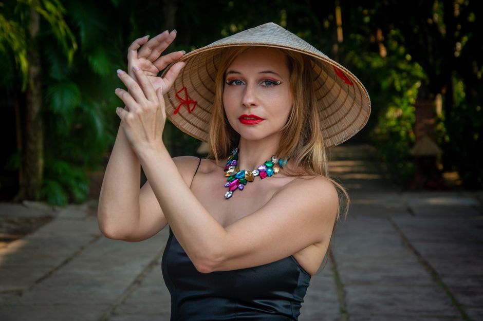 A stylish woman wearing a traditional Asian hat and vibrant necklace, outdoors in a serene setting.