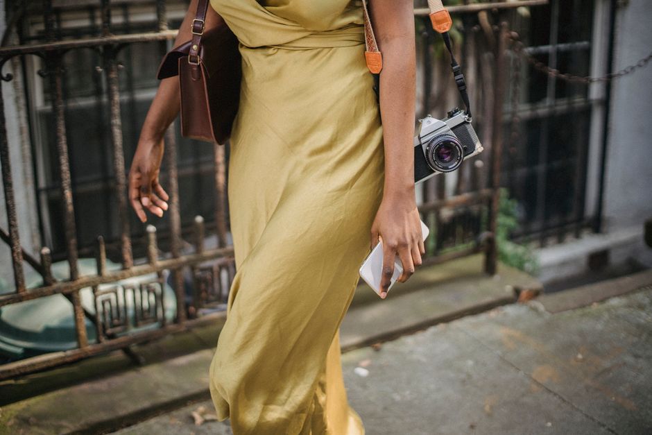 Fashionable woman in a yellow dress walking outdoors holding a vintage camera and smartphone.