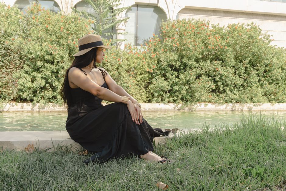 Woman in a black dress and sun hat sitting by a canal in Baku, enjoying a sunny day.
