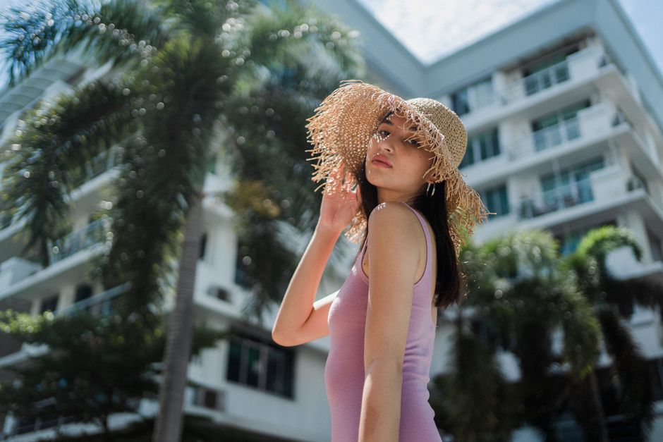 Side view of young feminine ethnic lady in top touching stylish straw hat and looking at camera while relaxing in modern resort during summer holidays