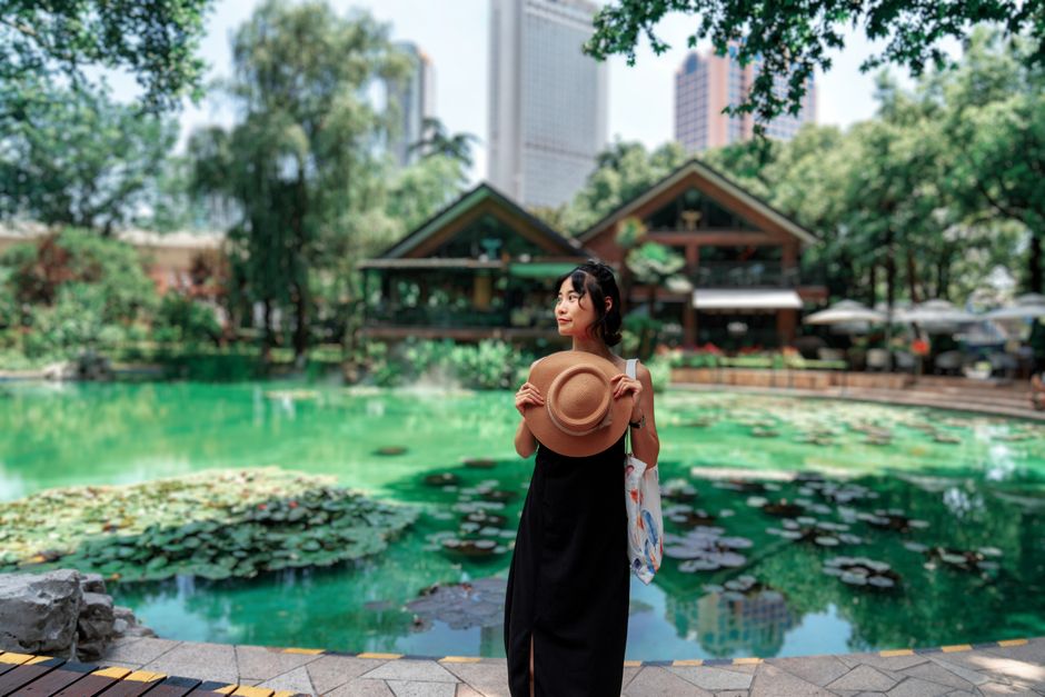 Asian woman holding hat by a pond in urban park setting, cityscape in background.