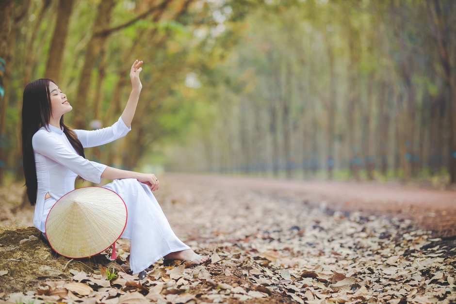 Young woman in Vietnamese ao dai sitting peacefully on a forest path holding a conical hat.