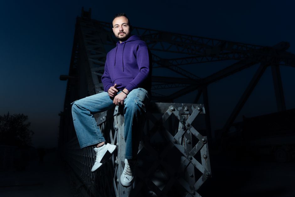 Casual man in purple hoodie sitting on bridge at night with a confident pose.