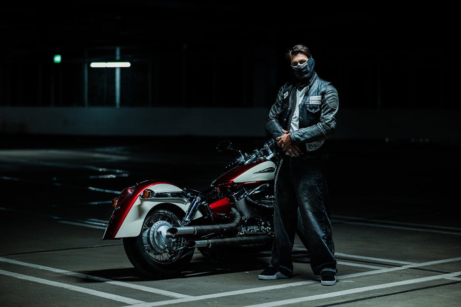 Motorcyclist in leather jacket standing by classic bike in dimly lit garage.