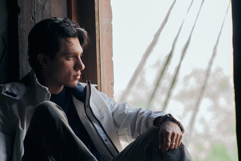 A young man gazes thoughtfully outside from a wooden window indoors.